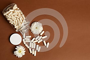 Open medicine bottle, scattered pills and flowers on brown background, flat lay. Space for text