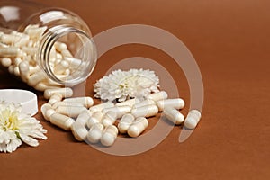 Open medicine bottle, scattered pills and flowers on brown background, space for text