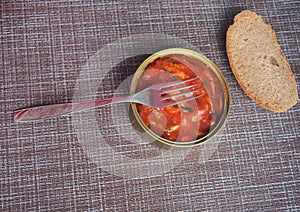 An open metal can of canned food, a fork, and a piece of bread lie on the brown tablecloth