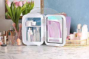 Open mini cosmetics refrigerator and skin care products on white marble table