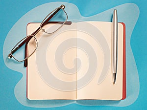 Open note, pen and eyeglasses top view on blue table