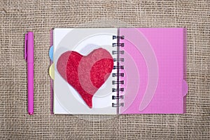 Open notebook with ballpoint pen and red heart of wool on a burlap background