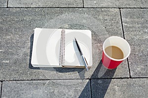 Open notebook with blank pages, pen and Cup of coffee on gray paving slab. Top view,