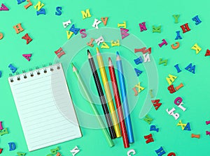 Open notebook with blank white sheets in line, colored wooden pencils on a green background with scattered wooden letters