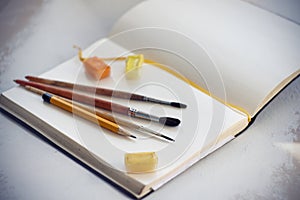 On an open notebook are brushes, pencil and watercolors in cuvettes