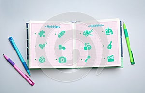 Open notebook with icons of hobbies and pens on light background, flat lay