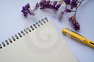 An open notebook for notes with a yellow pen on a white background, decorated by purple dried flowers. Shooting from above, flat