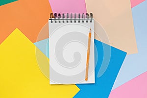 Open notebook with orange pencil on a multi colored paper background. Top view with copy space.