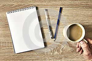 an open notebook with a pencil and a hand holding a cup of coffee on a wooden table, top view