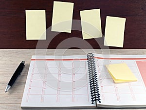 Open notebook with sticky notes and black ballpoint pen on wooden table and yellow blank notes on dark brown wooden wall