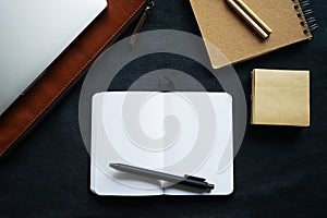Open notepad with black pen next to laptop case, notepad and pens on dark background