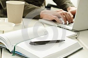 Open notepad with black pen on work-table. Paper cup of coffee, office stuff, laptop and working man on background