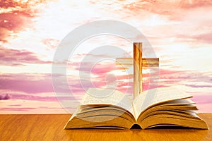 Open old bible on a wood table with blurred cross & colourful clouds and sky as background