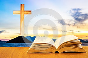 Open old bible on a wood table with blurred cross & colourful clouds and sky as background