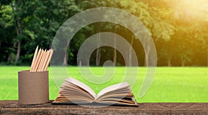 open old book and a pencil case on a wooden table. beautiful green lawn and forest background