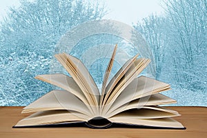 Open paper book on a background of a natural winter landscape on a wooden table, literary concept, close-up, copy space