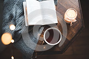 Open paper book with cup of coffee and burning candle on knit sweater on wooden table close up top view.