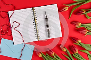 Open paper notebook, envelope and pen with red spring tulips flowers on red background.