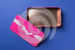 Open pink metal gift box with printed tape and bow on blue background , top view