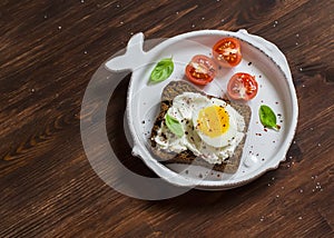 Open sandwich with feta cheese and boiled egg, tomatoes, and basil on a white plate on a wooden surface.