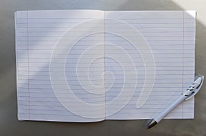 Open school notebook with pen on a gray desk with sun rays from the window. A blank, white sheet of notebook for writing.