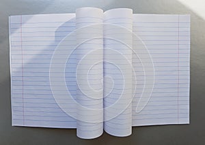 Open school notebook with pen on a gray desk with sun rays from the window. A blank, white sheet of notebook for writing.