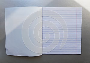 Open school notebook with pen on a gray desk with sun rays from the window. A blank, white sheet of notebook for writing.
