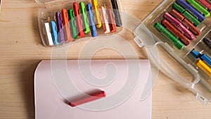 An open sketchbook, with a red crayon on top and several other colored crayons nearby, is in a box on a wooden table.