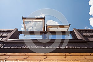 Open skylights mansard windows in wooden house with tile against blue sky.