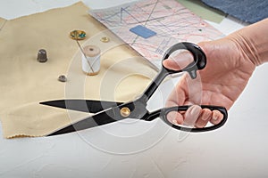Open tailor`s scissors in a female hand against the background of fabric and sewing accessories.