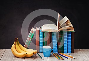 Open textbook, pile of books in colorful covers, bananas, pencils and cup of coffee on wooden table with green blackboard