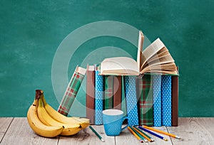 Open textbook, pile of books in colorful covers, bananas, pencils and cup of coffee on wooden table with green blackboard