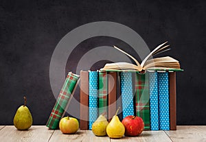 Open textbook, pile of books in colorful covers, pears and apples on wooden table with green blackboard background. Distance home
