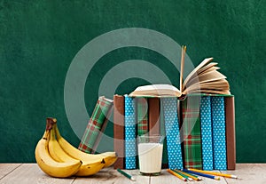 Open textbook, pile of books in colorful covers, pencils, bananas and a glass of milk on wooden table with green blackboard