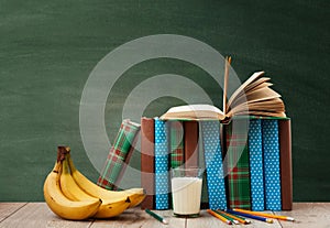 Open textbook  pile of books in colorful covers  pencils  bananas and a glass of milk on wooden table with green blackboard