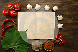 Open vintage book with berries, tomatoes, Chile peppers, spices and grape leaf on wooden background. Healthy vegetarian food.