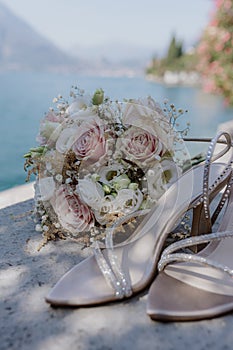 Open wedding shoes with rhinestones bouquet of bride lie against background of blue sky, sea.