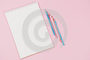 Open blank white notebook and light blue, pink, white colored pencils on pastel pink background. Top view, copy space