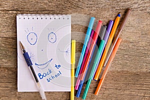 Open white notepad with drawing and inscription BACK TO SCHOOL with colorful felt-tip pens and ball pens on the wooden table