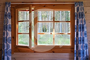 Open window with natural wooden frame and blue flower curtains. Morning forest view. Sunrise in the country