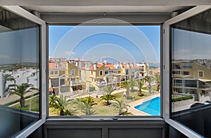 Through open window view to closed area of urbanization with planted palm trees on green lawn around swimming pool with blue water