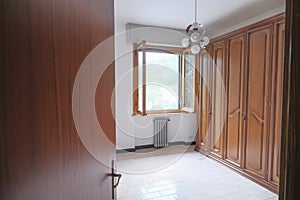 Open wooden door to a room with white walls window, open window, wooden wardrobe and chandelier on the ceiling. Home interior