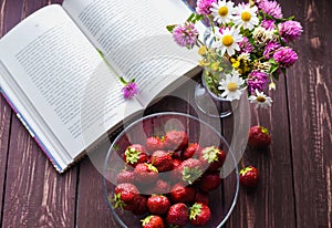 An opened book, a bowl with fresh mature appetizing strawbwrries and a bouquet of field flowers on a dark rustic wood