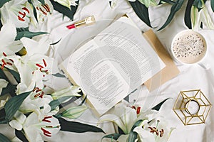 Opened book, coffee cup, candlestick, red lipstick and fresh flowers on the white bed. Flat lay, top view