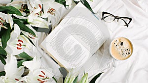 Opened book, coffee cup, glasses and fresh flowers on the white bed. Flat lay, top view