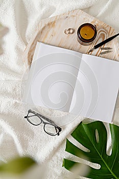 Opened book, glasses, candle and flowers top view on white bed. Mock up design