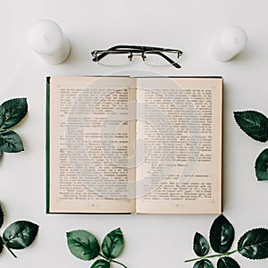 Opened book, glasses, green leaves on white background. Flat Lay