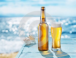 Opened bottle of beer and beer glass with condensation on the wooden table. Blurred sparkling sea at the background. Summer vibes