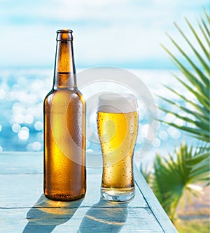 Opened bottle of beer and beer glass with condensation on the wooden table. Blurred sparkling sea at the background. Summer vibes