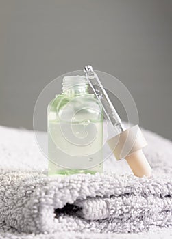 Opened cosmetic bottle with green serum on folded grey bath towel close up